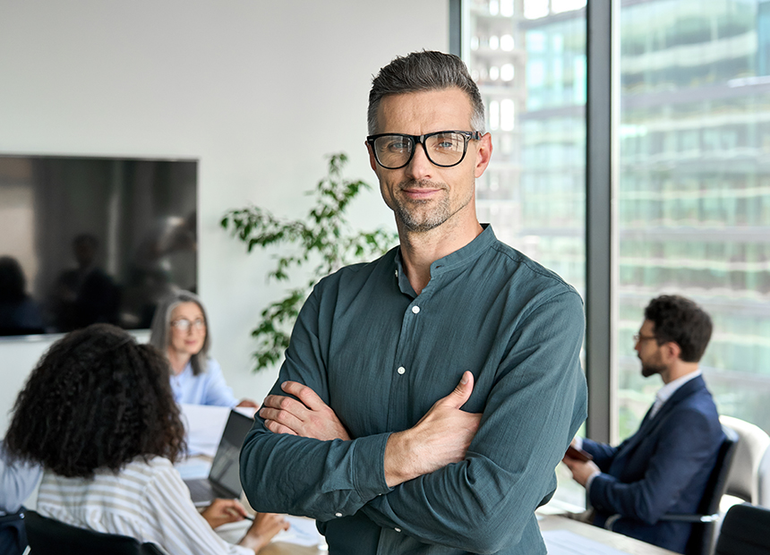 Smiling confident mature businessman leader looking at camera standing in office at team meeting. Male corporate leader ceo executive manager wearing glasses posing for business portrait arms folded.